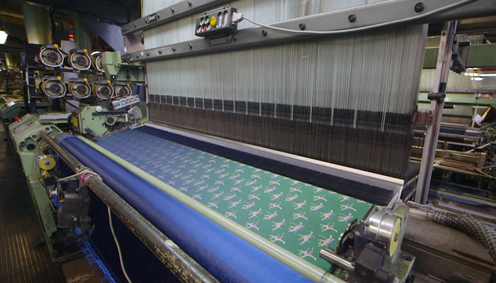 A textile machine is weaving fabric with a repeating airplane pattern. Multiple spools of thread are visible on the side, and the fabric is being produced in a busy industrial setting.