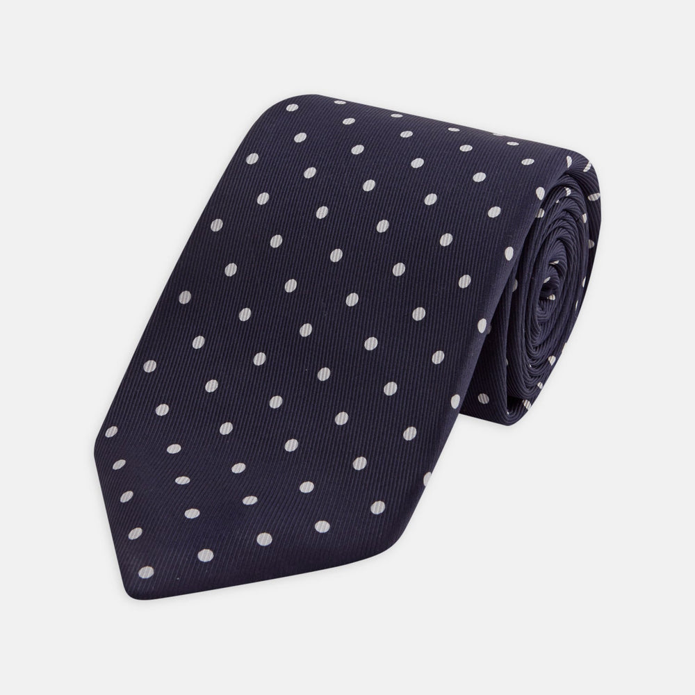 The Seven-Fold Navy Churchill Spot Silk Tie by Turnbull & Asser features evenly spaced white polka dots on navy silk—a luxury neckwear piece rolled up and displayed on a white background.