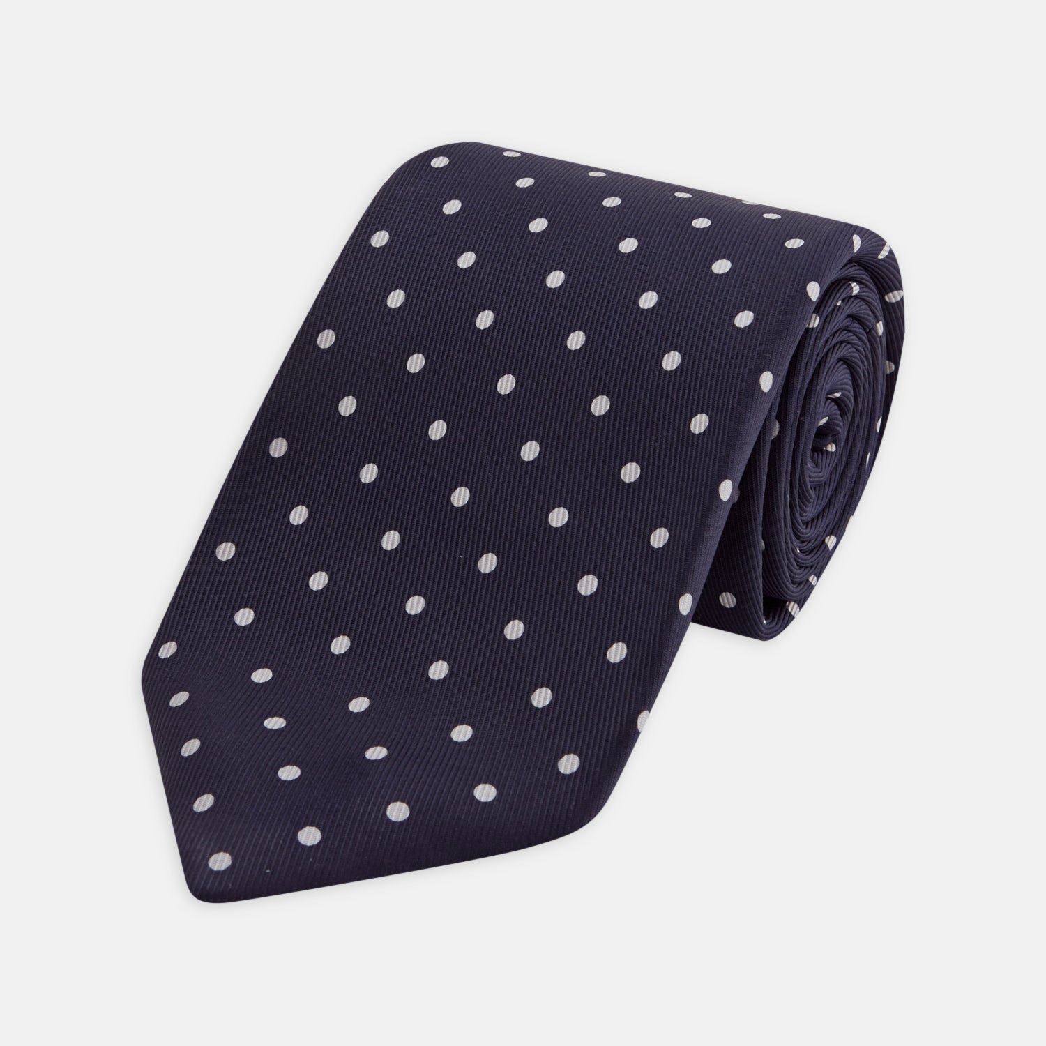 View 1 of product The Seven-Fold Navy Churchill Spot Silk Tie by Turnbull & Asser features evenly spaced white polka dots on navy silk—a luxury neckwear piece rolled up and displayed on a white background.
