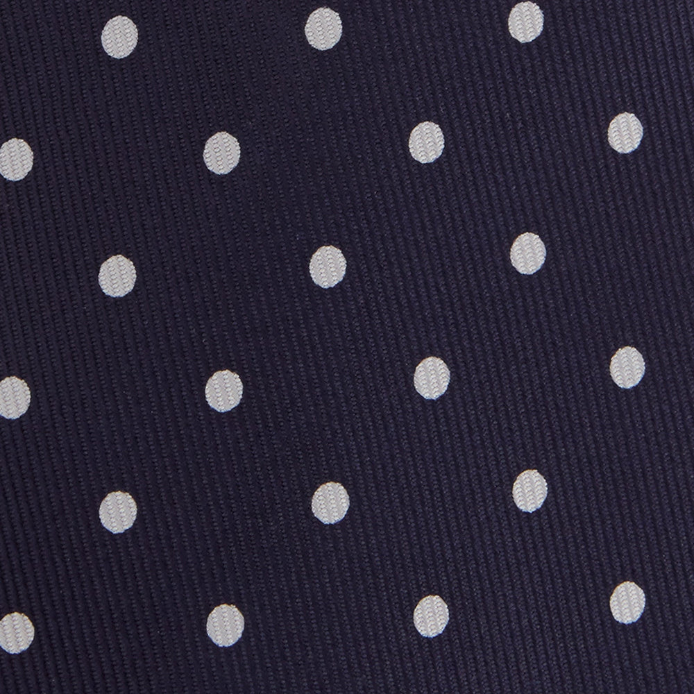 The Seven-Fold Navy Churchill Spot Silk Tie by Turnbull & Asser features evenly spaced white polka dots on navy silk—a luxury neckwear piece rolled up and displayed on a white background.