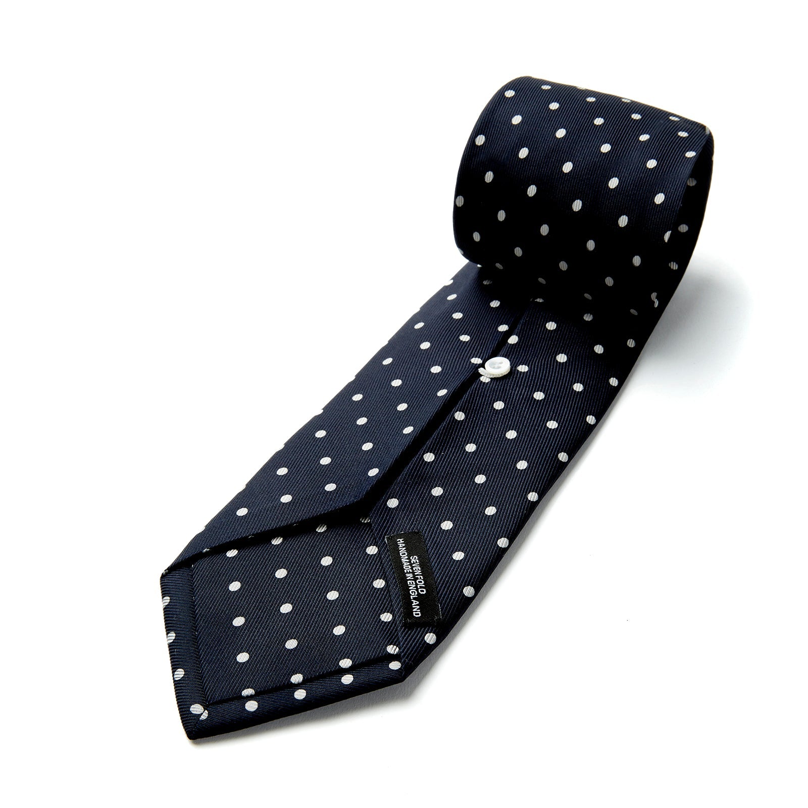 View 4 of product The Seven-Fold Navy Churchill Spot Silk Tie by Turnbull & Asser features evenly spaced white polka dots on navy silk—a luxury neckwear piece rolled up and displayed on a white background.
