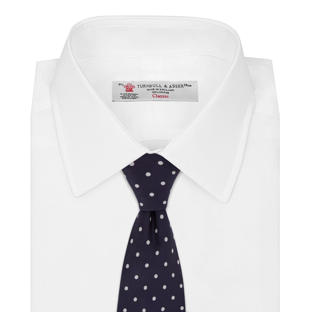The Seven-Fold Navy Churchill Spot Silk Tie by Turnbull & Asser features evenly spaced white polka dots on navy silk—a luxury neckwear piece rolled up and displayed on a white background.