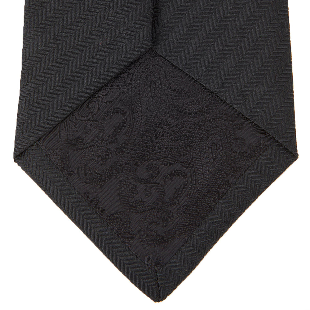 The Turnbull & Asser Black Herringbone Silk Tie is rolled up and displayed on a plain white background, showcasing its classic style and subtle chevron pattern.