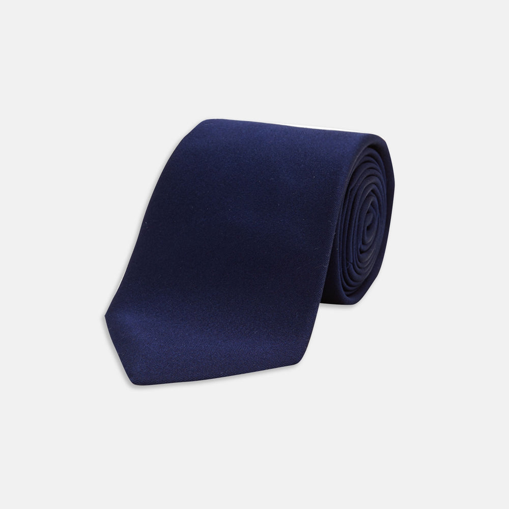 The Turnbull & Asser Seven-Fold Navy Plain Satin Silk Tie is neatly rolled and showcased against a plain light gray background.