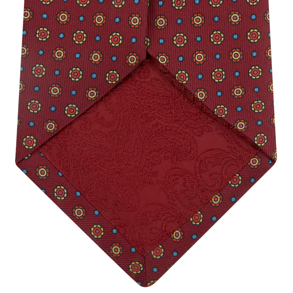 The Turnbull & Asser "The Great Gatsby" Burgundy Printed Silk Tie, from the Legends collection, showcases small circular floral motifs in yellow, blue, and green. Rolled with the tip forward, it is displayed against a white background.