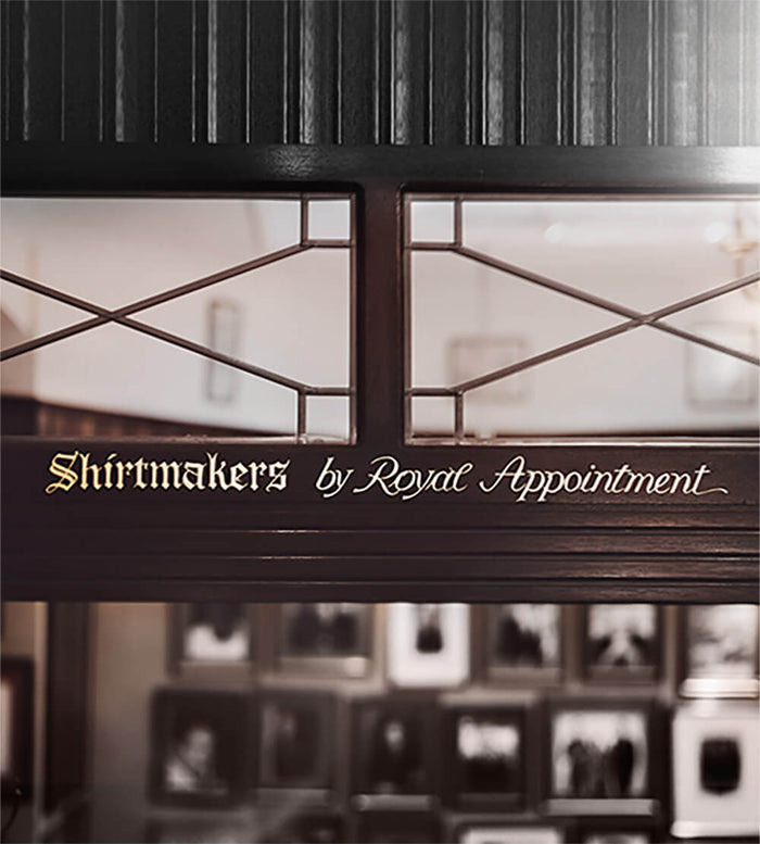 A shopfront with dark wood and glass panels displays the gold-lettered sign: Shirtmakers by Royal Appointment. Framed photos are visible inside on the back wall.