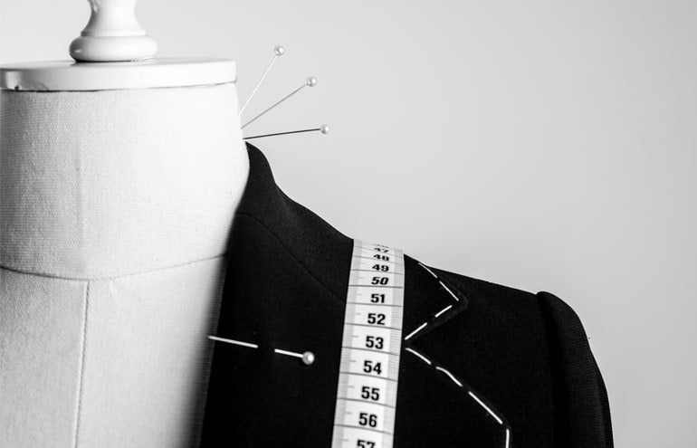A close-up of a dress form showcasing Turnbull & Asser's Clothing Alterations (Bradford): a partially tailored black jacket with white stitching, a measuring tape over the shoulder, and pins marking adjustments.