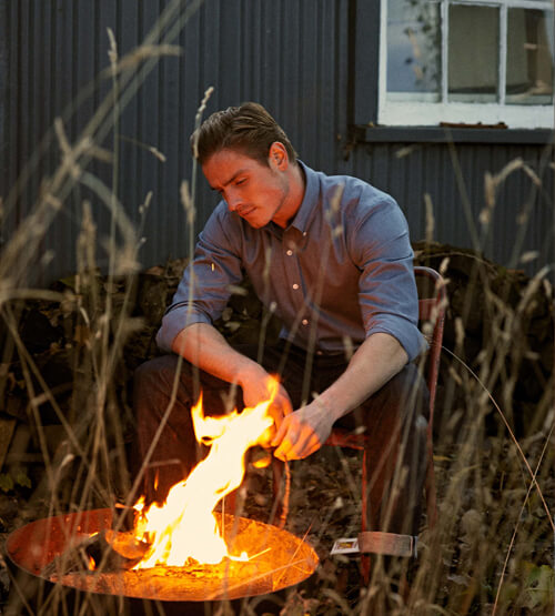 A man in a blue shirt sits on a chair outdoors, tending to a fire in a fire pit. Tall dry grass is in the foreground, and a dark building with a window is in the background.