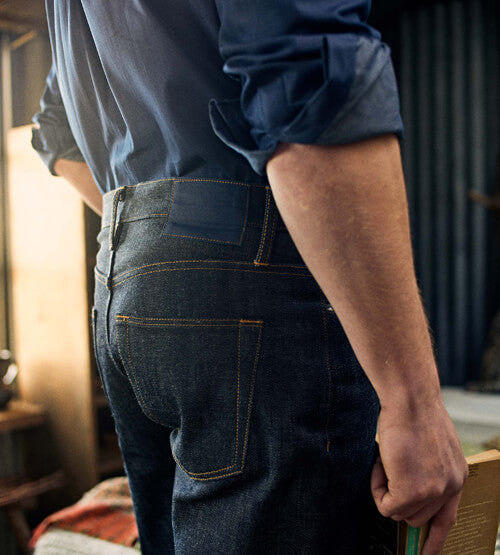 A person wearing a blue shirt and dark denim jeans stands indoors, holding a book by their side. The photo is taken from behind, focusing on the jeans and the person’s left arm.