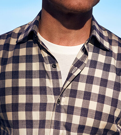A close-up of a person wearing a navy and white checkered button-up shirt over a plain white T-shirt, with sunlight illuminating the scene. The persons face is partially visible.