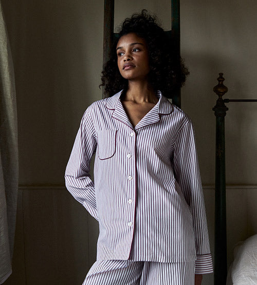 A woman with curly hair stands indoors wearing a matching striped pajama set with a collared shirt and pants, looking off to the side with a calm expression.