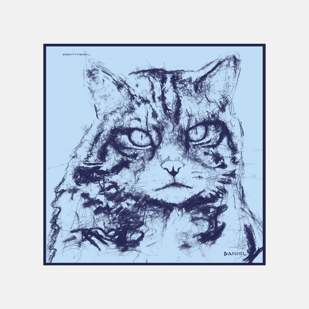 The Turnbull & Asser DANIEL PERKIN BLUE WILDCAT POCKET SQUARE features a blue-toned sketch of a tabby cat’s face with vivid eyes and fine fur detail on a pale blue background, finished with “DANISL” in the lower right corner.