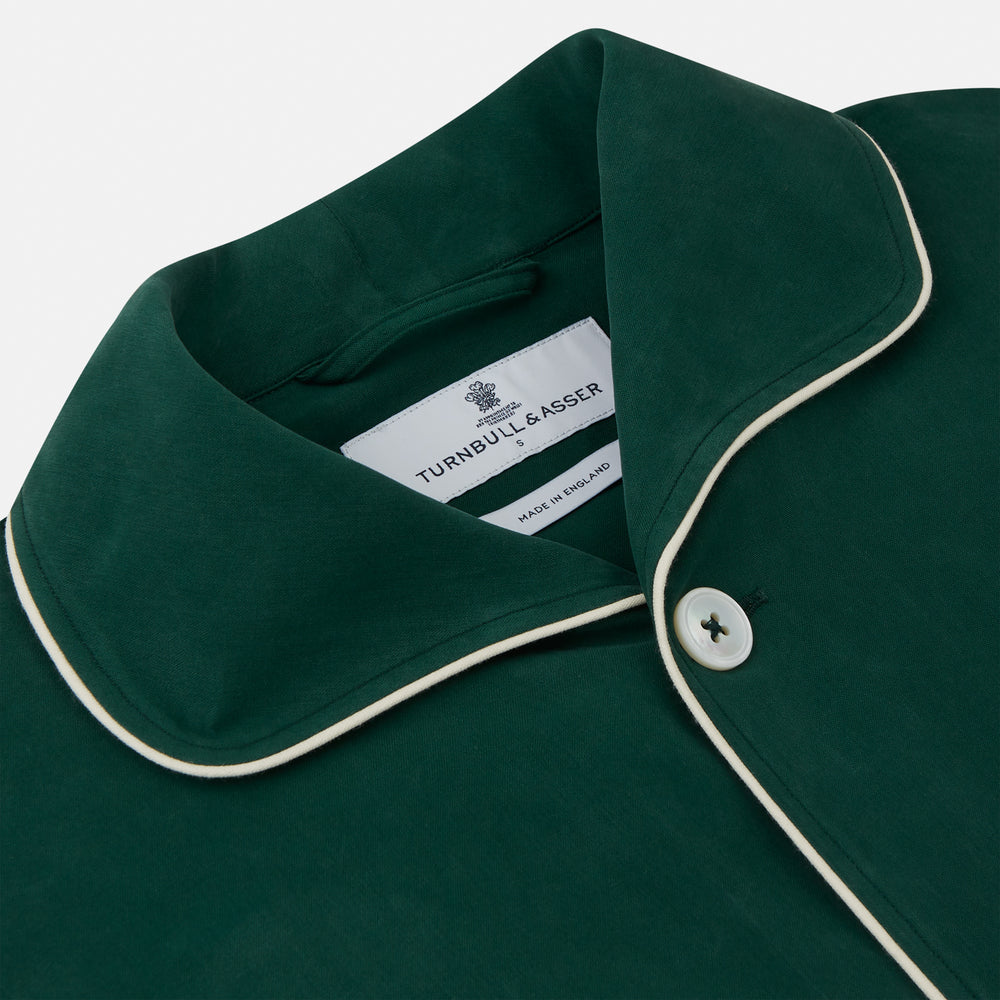 The Turnbull & Asser Dark Green Silk Pyjama Set features a long-sleeve button-up top with white piping, chest pocket, and matching pants, all neatly folded on a plain white background.