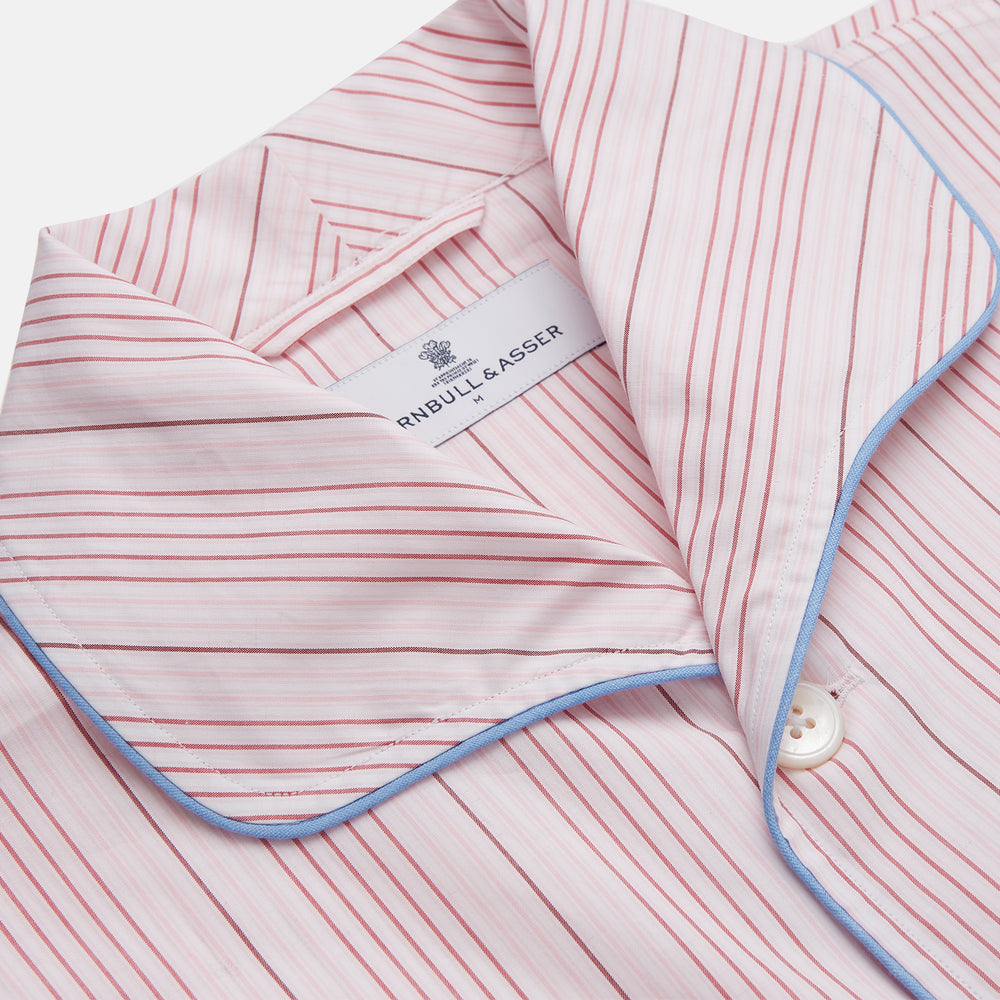 The Turnbull & Asser Pink And White Stripe Twill Cotton Pyjama Set features long sleeves and pants with a curved collar and blue trim on the cuffs, pocket, and hem—a classic gent’s style shown on a plain white background.