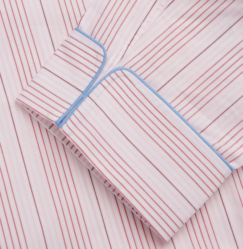 The Turnbull & Asser Pink And White Stripe Twill Cotton Pyjama Set features long sleeves and pants with a curved collar and blue trim on the cuffs, pocket, and hem—a classic gent’s style shown on a plain white background.