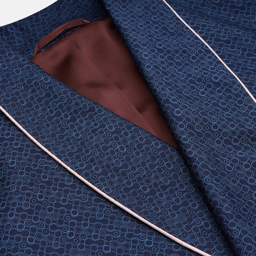 The Turnbull & Asser Blue Multi Circle Silk Jacquard Gown features a subtle geometric pattern, white piping, a chest pocket, and a matching waist belt, all showcased against a white background.