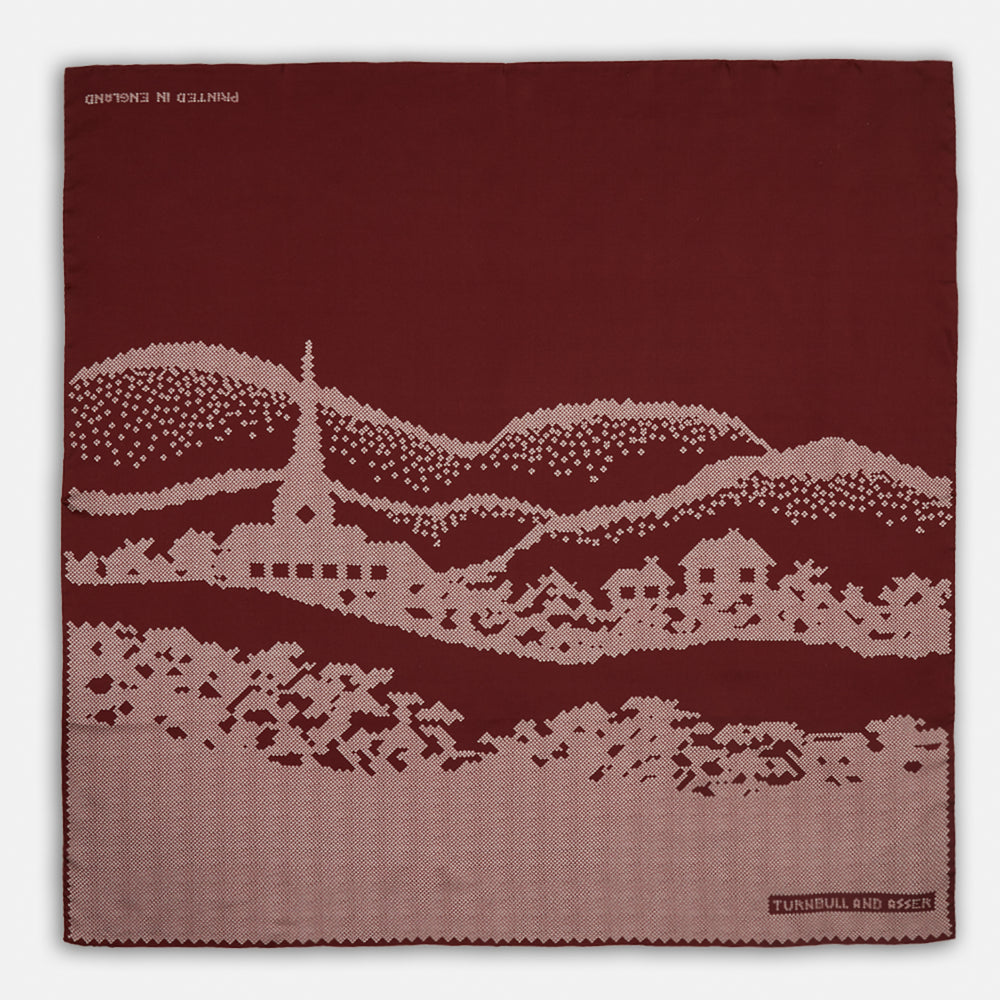 The Burgundy Snowscape Motif Silk Extra Large Scarf by Turnbull & Asser features a hand-rolled edge and a pink cross-stitch village scene—houses, church, hills—printed in England. A refined winter accessory.