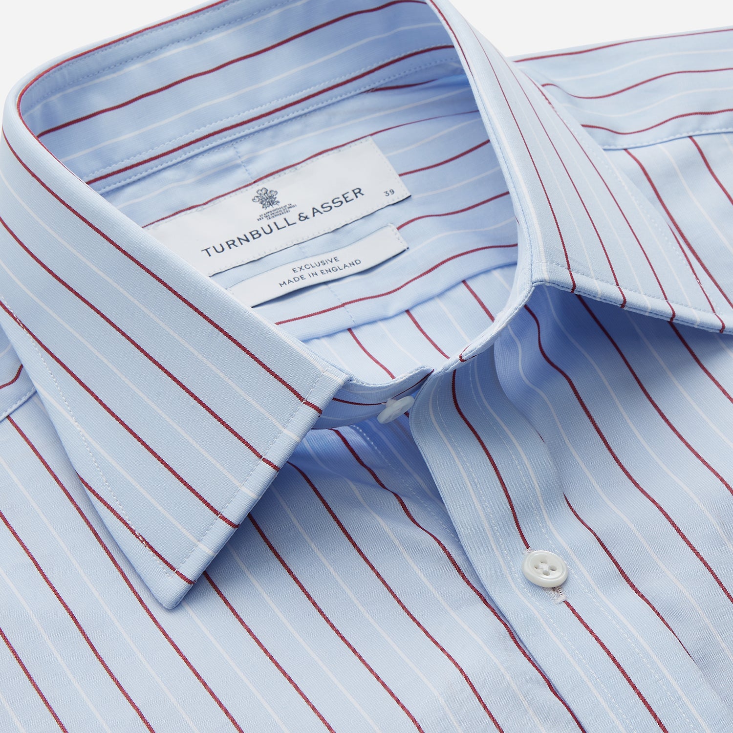 View 2 of product The Turnbull & Asser Burgundy Stripe Regular Fit Shirt features a T&A collar, double cuffs, and white buttons. Made from light blue Italian cotton poplin with vertical burgundy and white pinstripes. Laid flat on a white background.
