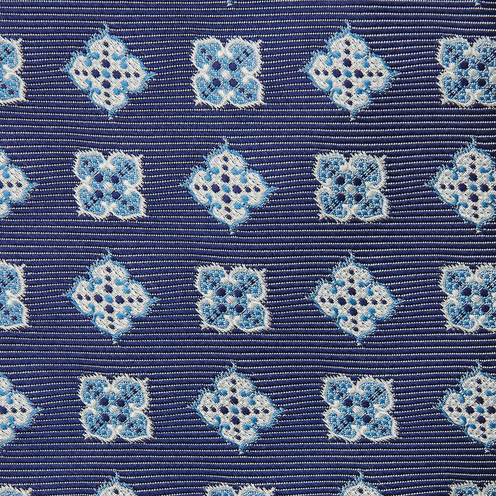The Turnbull & Asser Blue Geometric Jacquard Tie is a rolled-up silk tie showcasing a repeating light blue and white geometric floral pattern with dotted accents, set against a plain light background.