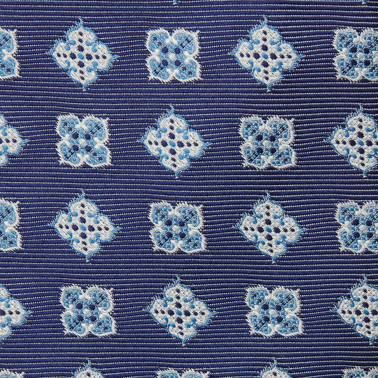 View 3 of product The Turnbull & Asser Blue Geometric Jacquard Tie is a rolled-up silk tie showcasing a repeating light blue and white geometric floral pattern with dotted accents, set against a plain light background.
