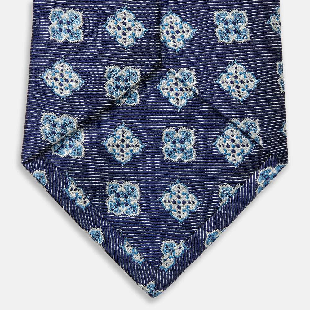 The Turnbull & Asser Blue Geometric Jacquard Tie is a rolled-up silk tie showcasing a repeating light blue and white geometric floral pattern with dotted accents, set against a plain light background.