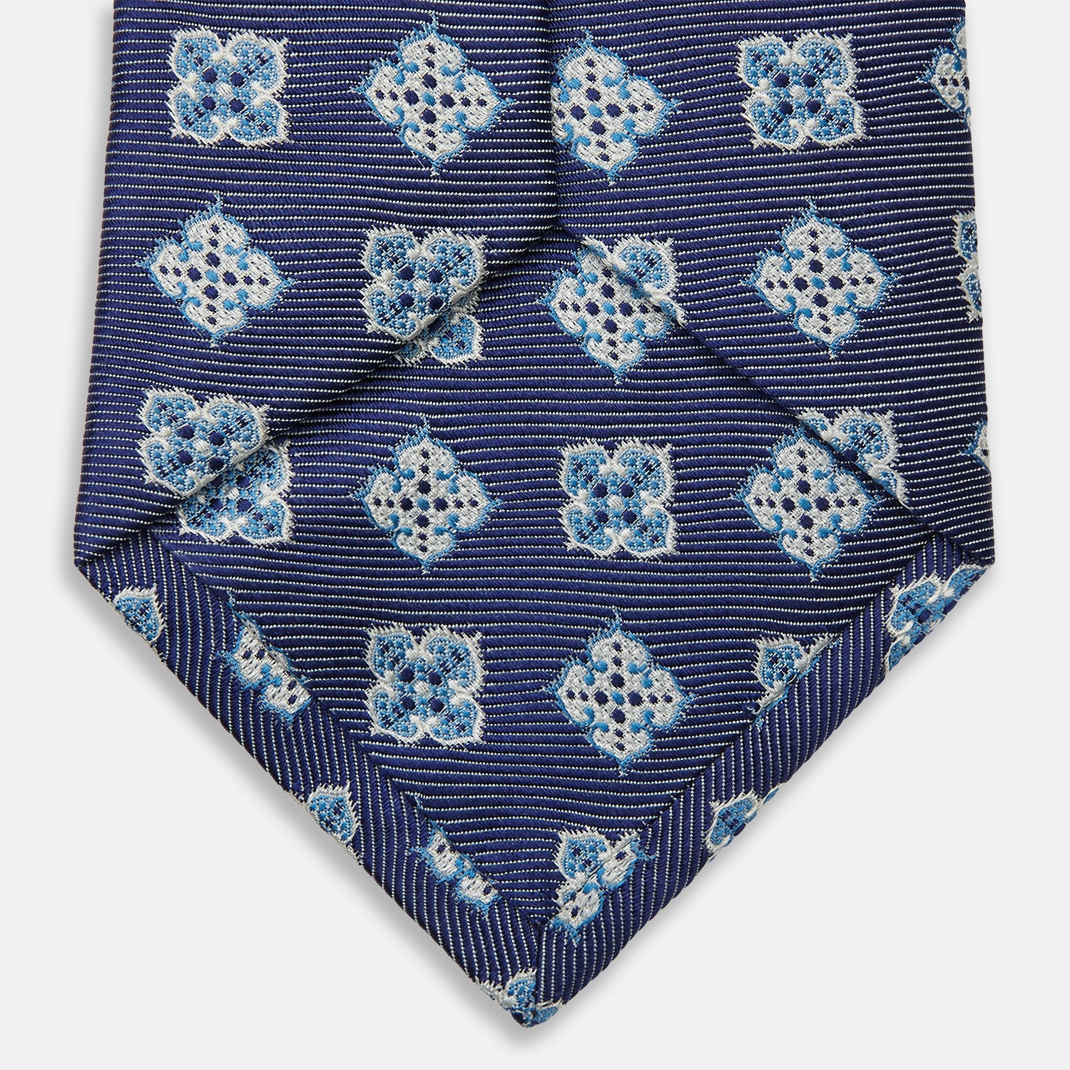 View 4 of product The Turnbull & Asser Blue Geometric Jacquard Tie is a rolled-up silk tie showcasing a repeating light blue and white geometric floral pattern with dotted accents, set against a plain light background.
