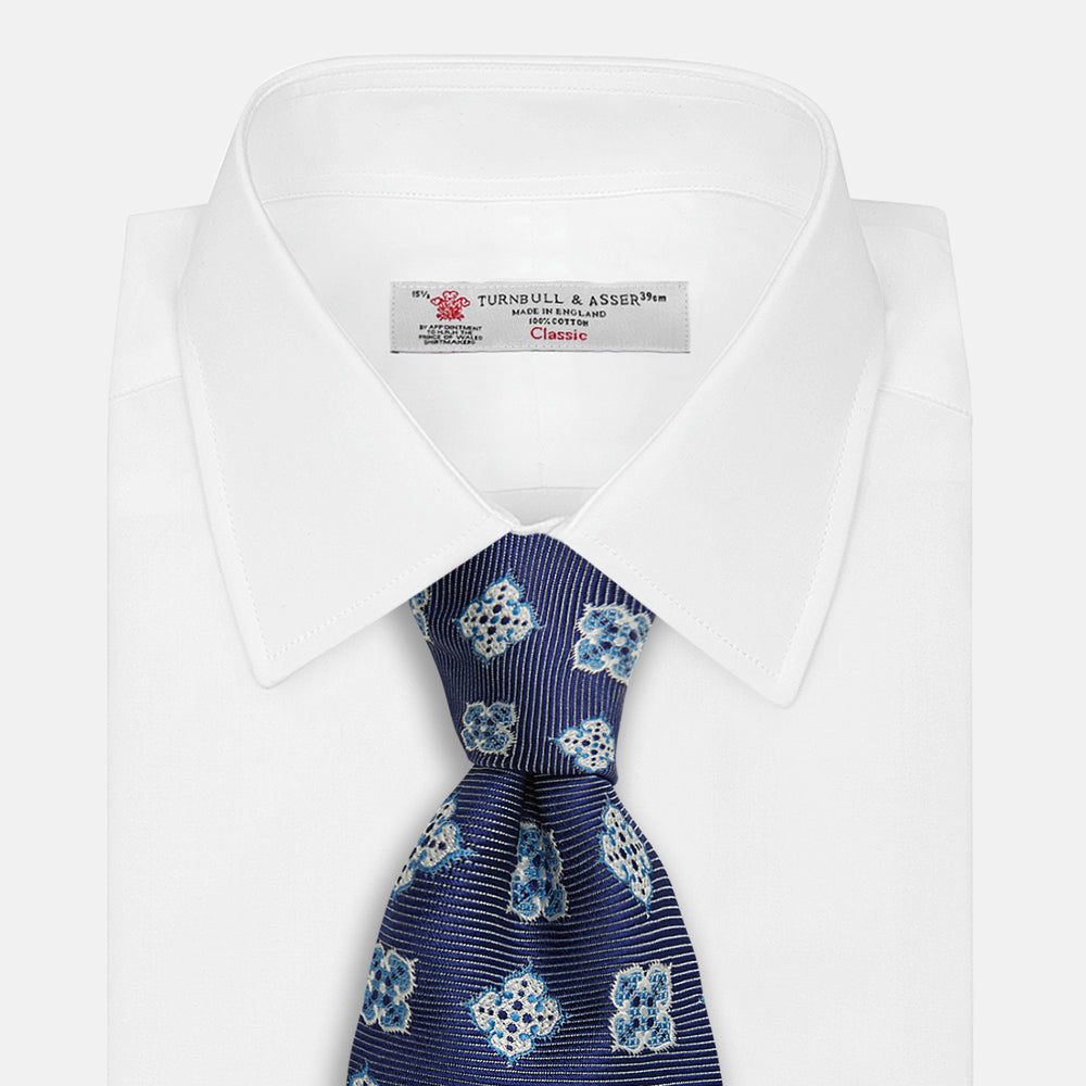 The Turnbull & Asser Blue Geometric Jacquard Tie is a rolled-up silk tie showcasing a repeating light blue and white geometric floral pattern with dotted accents, set against a plain light background.
