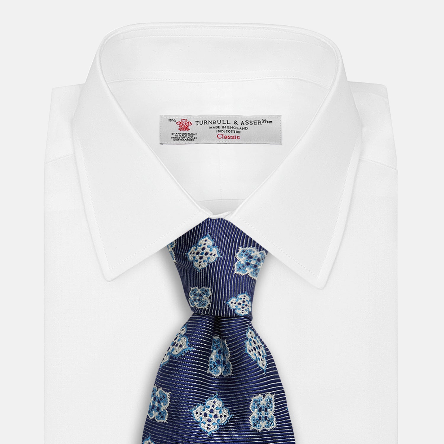 View 2 of product The Turnbull & Asser Blue Geometric Jacquard Tie is a rolled-up silk tie showcasing a repeating light blue and white geometric floral pattern with dotted accents, set against a plain light background.