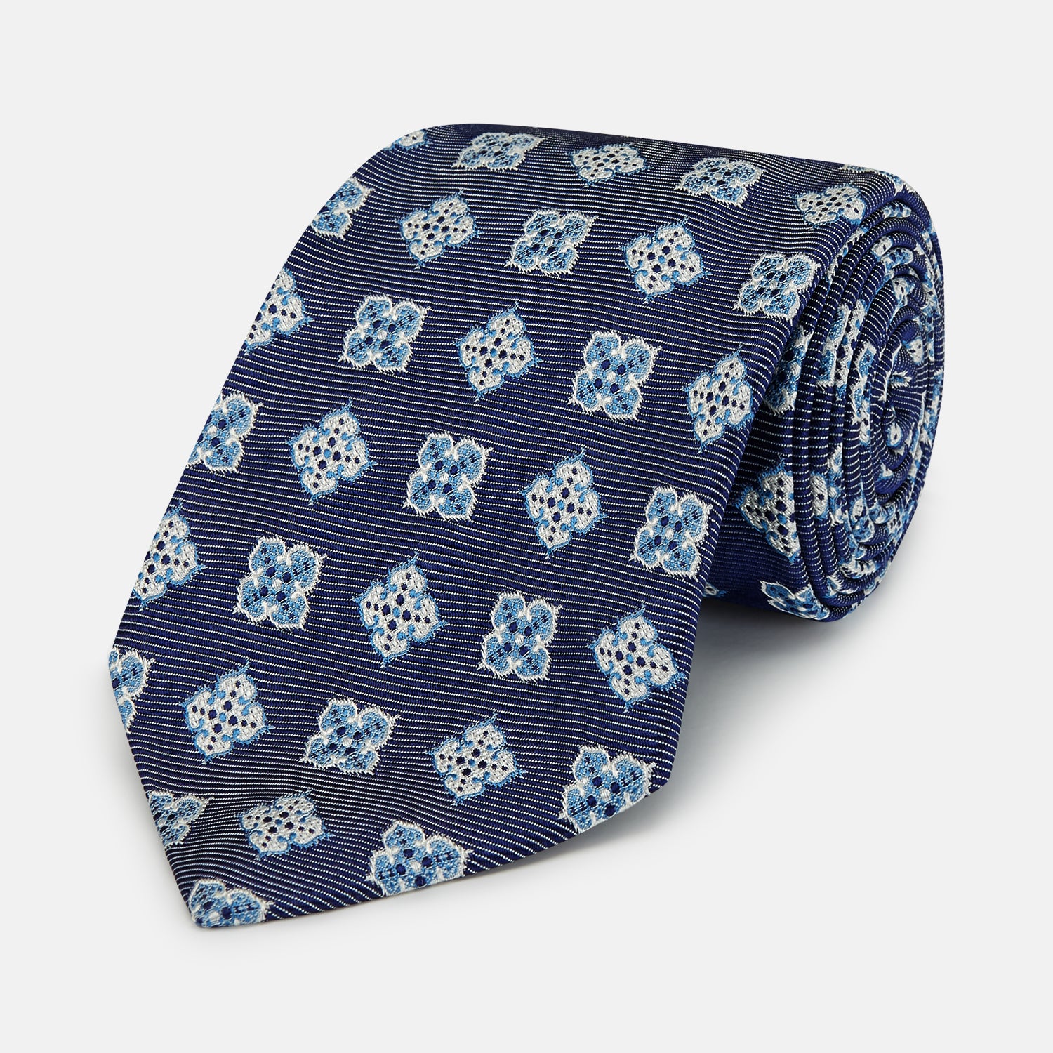 View 1 of product The Turnbull & Asser Blue Geometric Jacquard Tie is a rolled-up silk tie showcasing a repeating light blue and white geometric floral pattern with dotted accents, set against a plain light background.