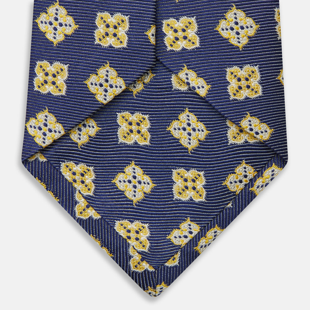 The Turnbull & Asser Yellow Geometric Jacquard Tie in navy features a repeating pattern of yellow geometric shapes with blue accents, presented rolled up on a white background.