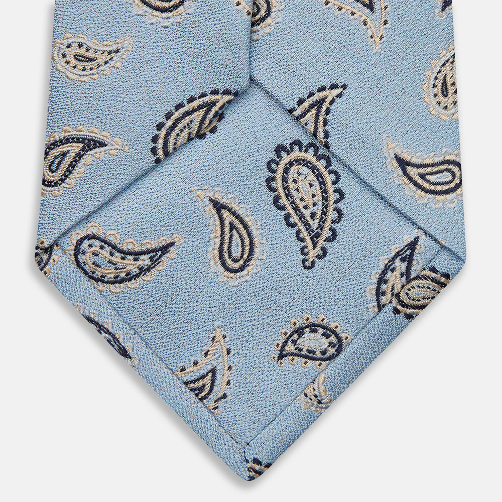 The Turnbull & Asser Sky Blue Renoir Paisley Tie, crafted in silk with a repeating navy and beige pattern, is shown rolled up with its pointed end forward on a plain white background.