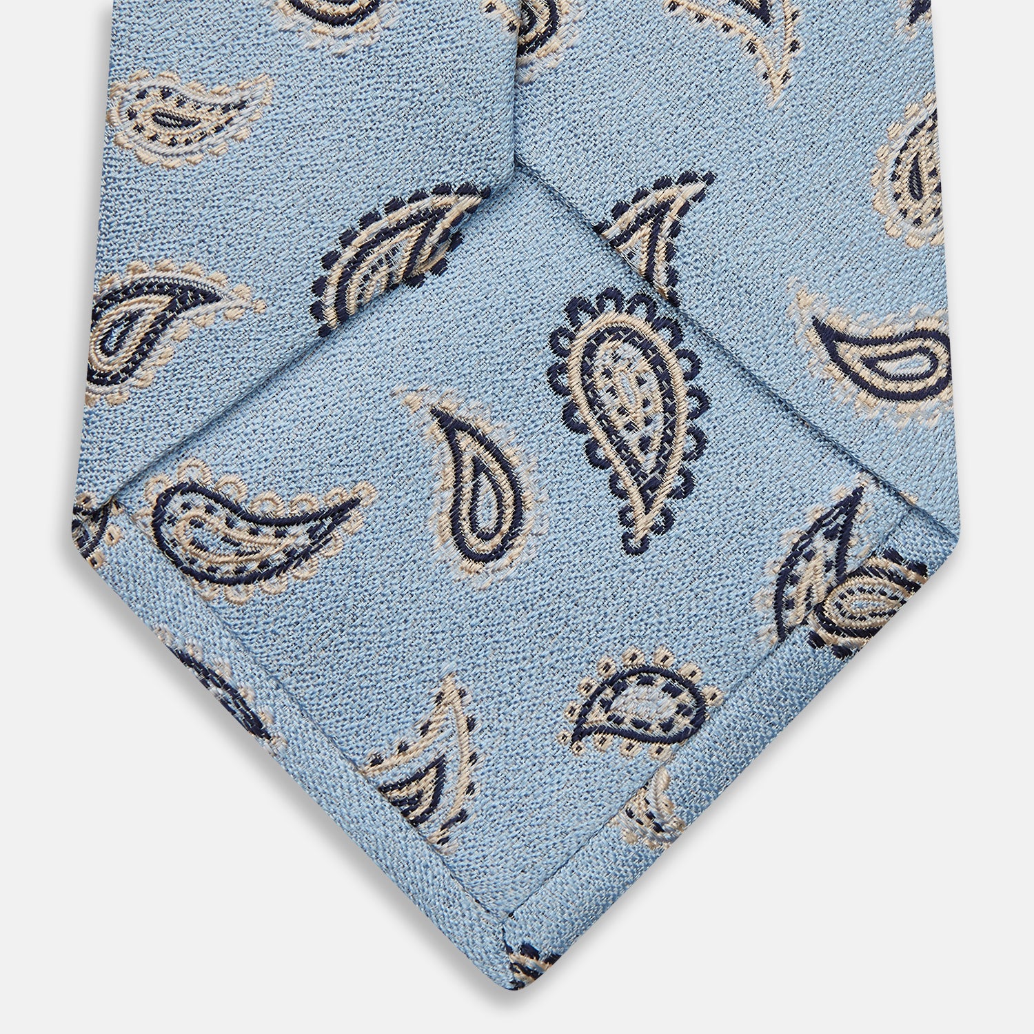 View 4 of product The Turnbull & Asser Sky Blue Renoir Paisley Tie, crafted in silk with a repeating navy and beige pattern, is shown rolled up with its pointed end forward on a plain white background.