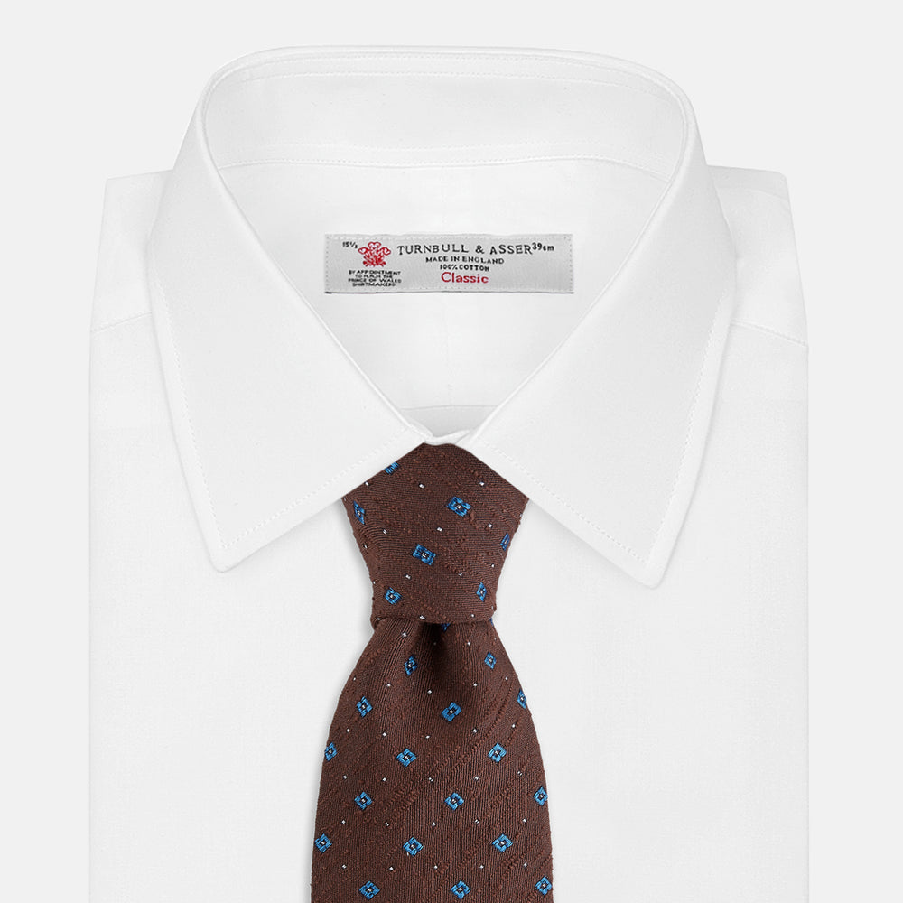 The Turnbull & Asser Brown Square Silk Tie, featuring a jacquard pattern of small blue squares and dots, is shown rolled up on a plain white background.