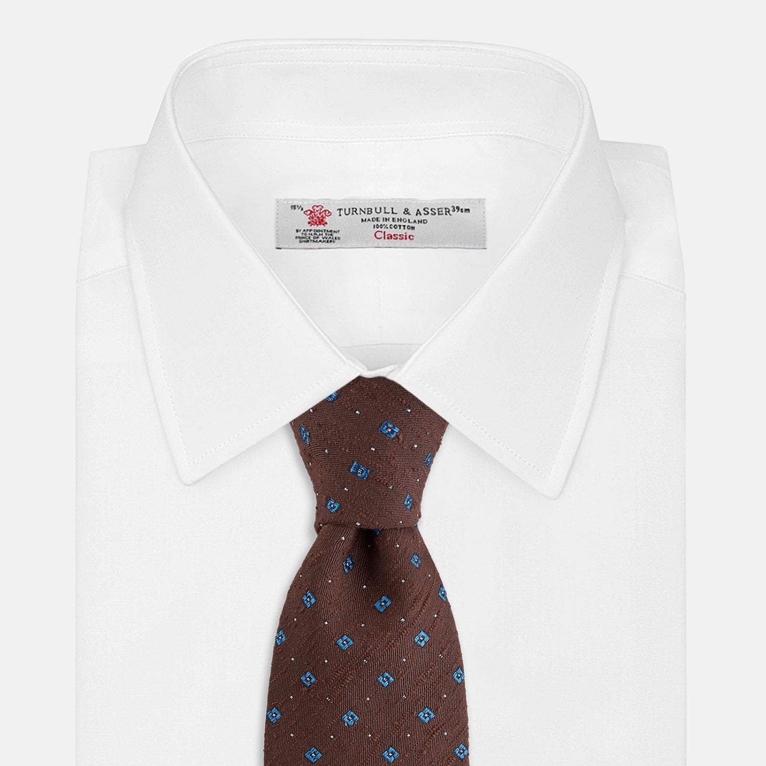 View 2 of product The Turnbull & Asser Brown Square Silk Tie, featuring a jacquard pattern of small blue squares and dots, is shown rolled up on a plain white background.