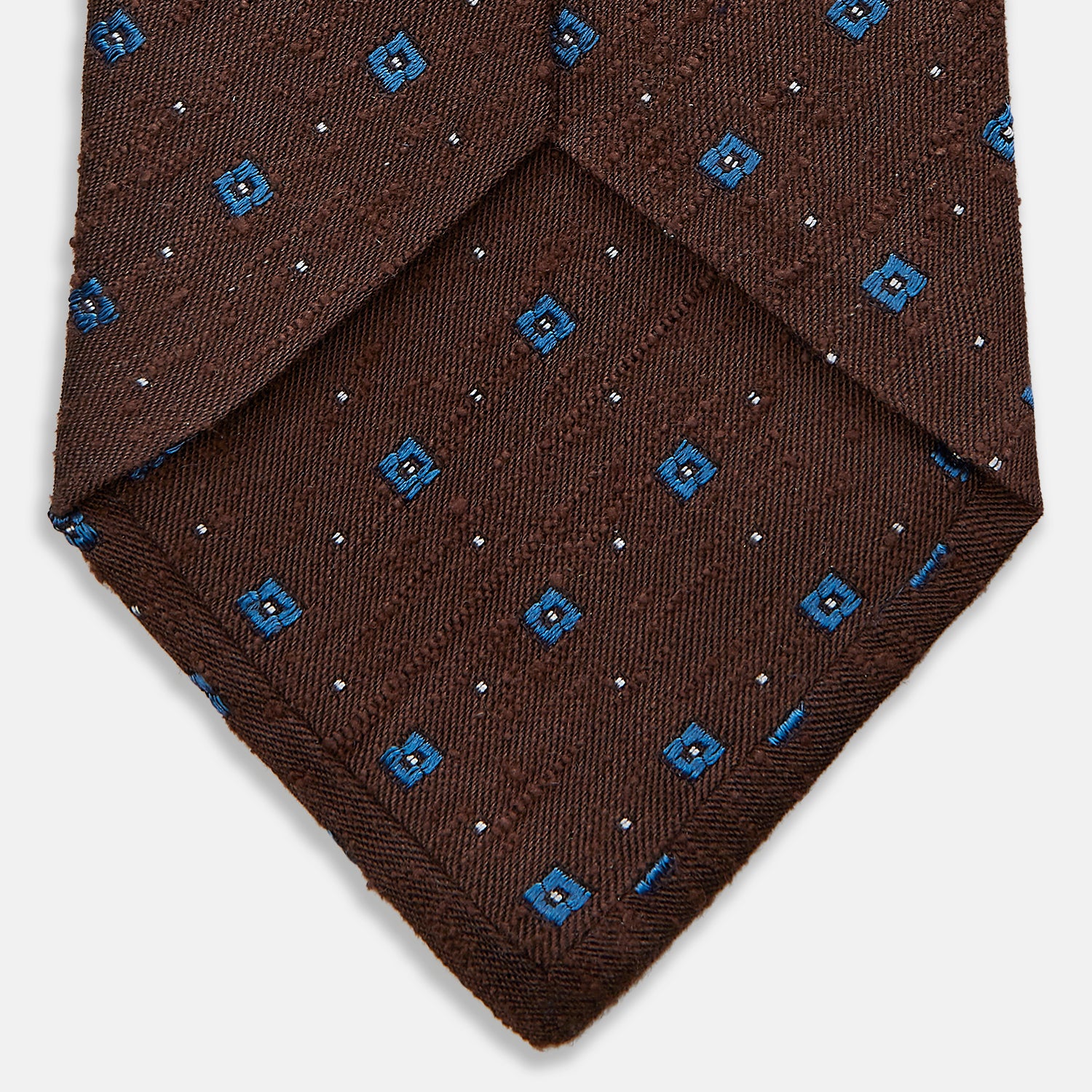 View 4 of product The Turnbull & Asser Brown Square Silk Tie, featuring a jacquard pattern of small blue squares and dots, is shown rolled up on a plain white background.