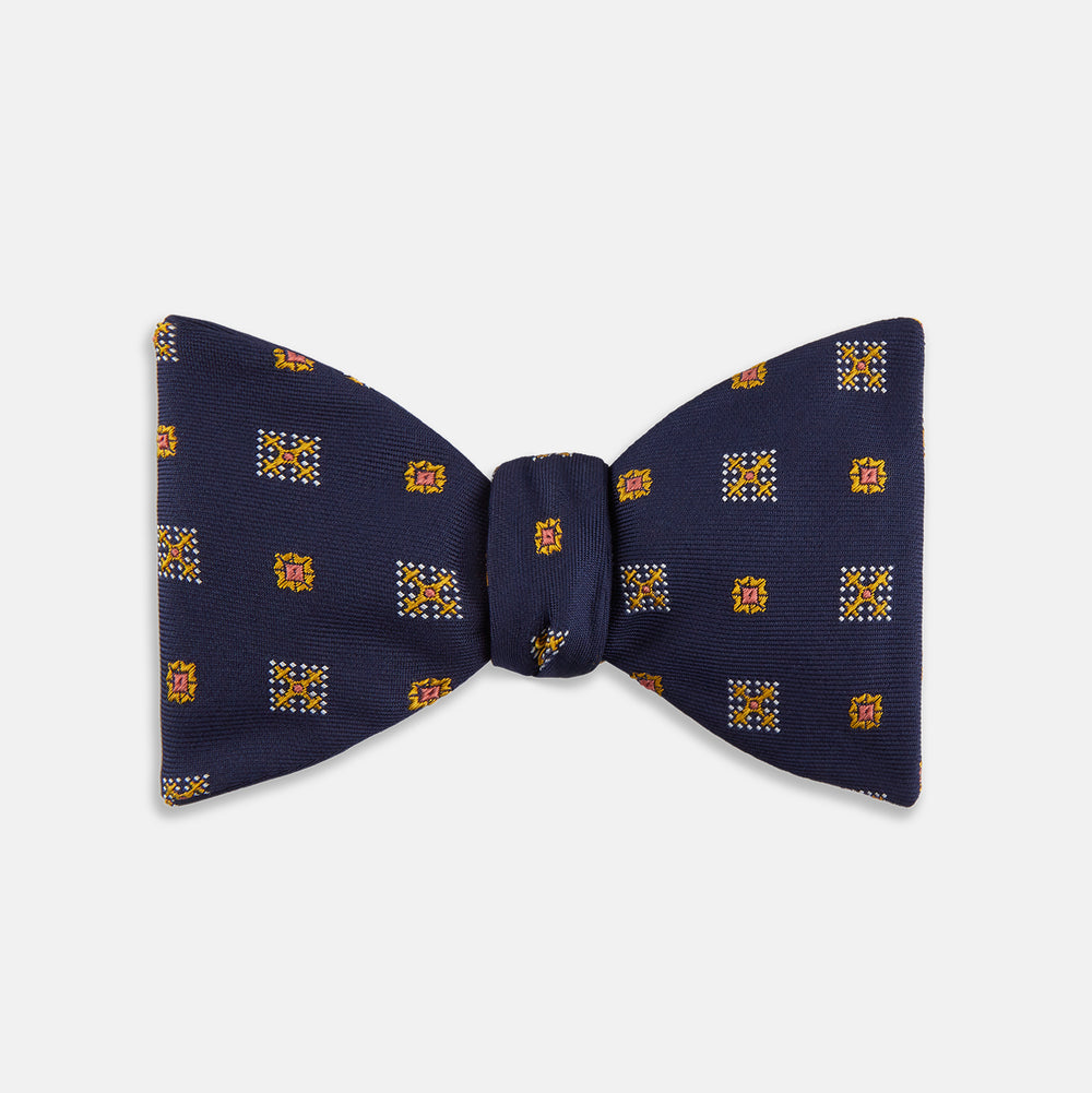 The Yellow Shields Bow Tie by Turnbull & Asser features a navy blue base with small yellow, orange, and white geometric squares, ideal for formal occasions and shown against a white background.