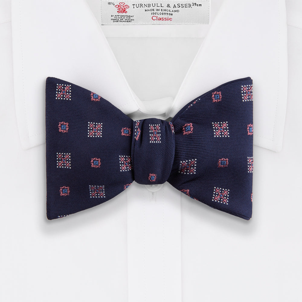 The Turnbull & Asser Pink Shields Bow Tie features a pink pattern with shield motifs, ideal for formal occasions and displayed on a plain white background.