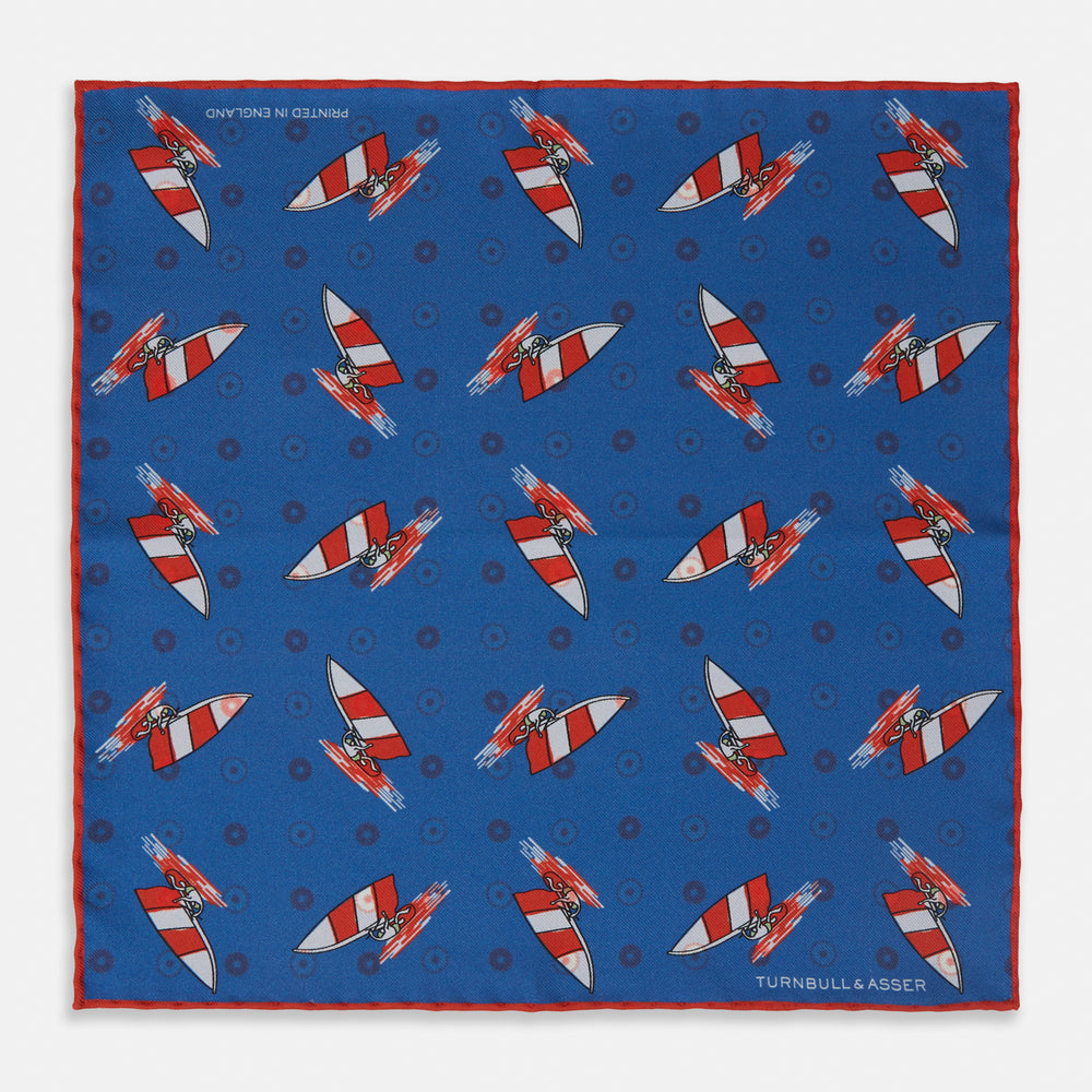 The Turnbull & Asser Blue Windsurfer Double-Faced Silk Print Pocket Square features a blue silk design with colorful windsurfers and vivid red edges, finished with the Turnbull & Asser logo in one corner.