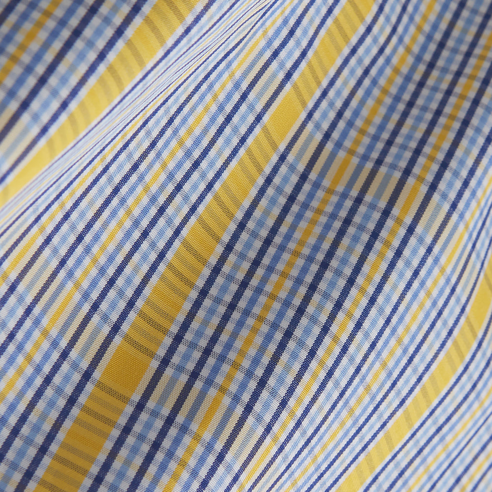 A man stands against a light background, wearing a Turnbull & Asser Yellow and Blue Fancy Check Regular Fit Shirt with T&A Collar, a patterned tie, dark dress pants, and black shoes. He faces forward with hands in pockets and a neutral expression.