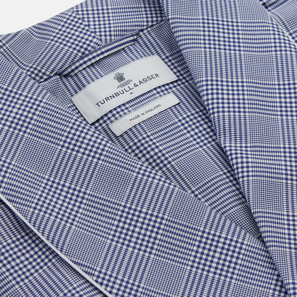 The Turnbull & Asser Navy Check Cotton Gown features long sleeves, white piping, a shawl collar, chest pocket, and matching waist belt, all in a blue plaid pattern. Shown against a plain white background.