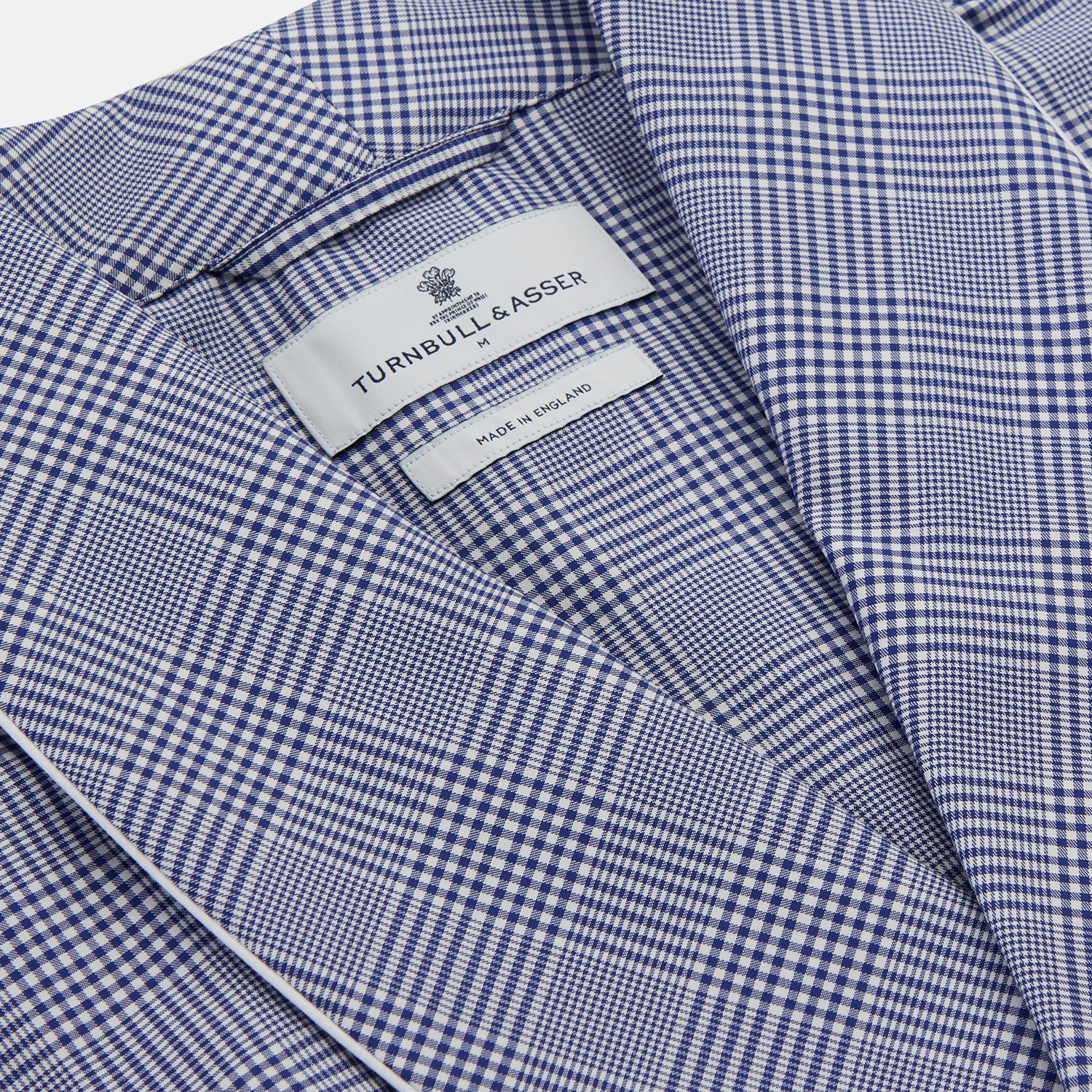 View 2 of product The Turnbull & Asser Navy Check Cotton Gown features long sleeves, white piping, a shawl collar, chest pocket, and matching waist belt, all in a blue plaid pattern. Shown against a plain white background.