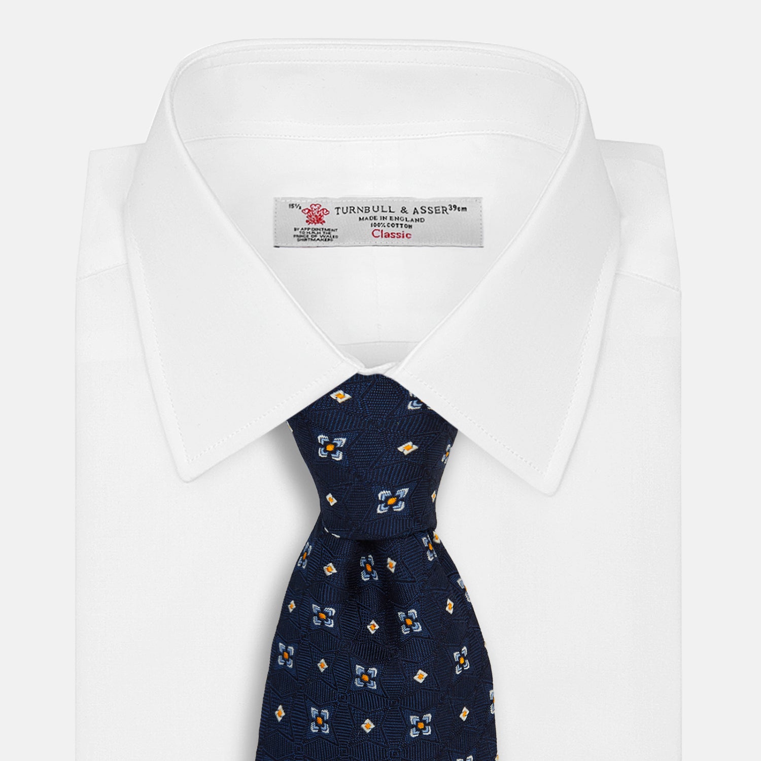 View 2 of product The Turnbull & Asser Navy Floral Emblem Tie displays a repeating pattern of small light blue, white, and yellow floral and diamond motifs, highlighting refined elegance, shown rolled up on a white background.