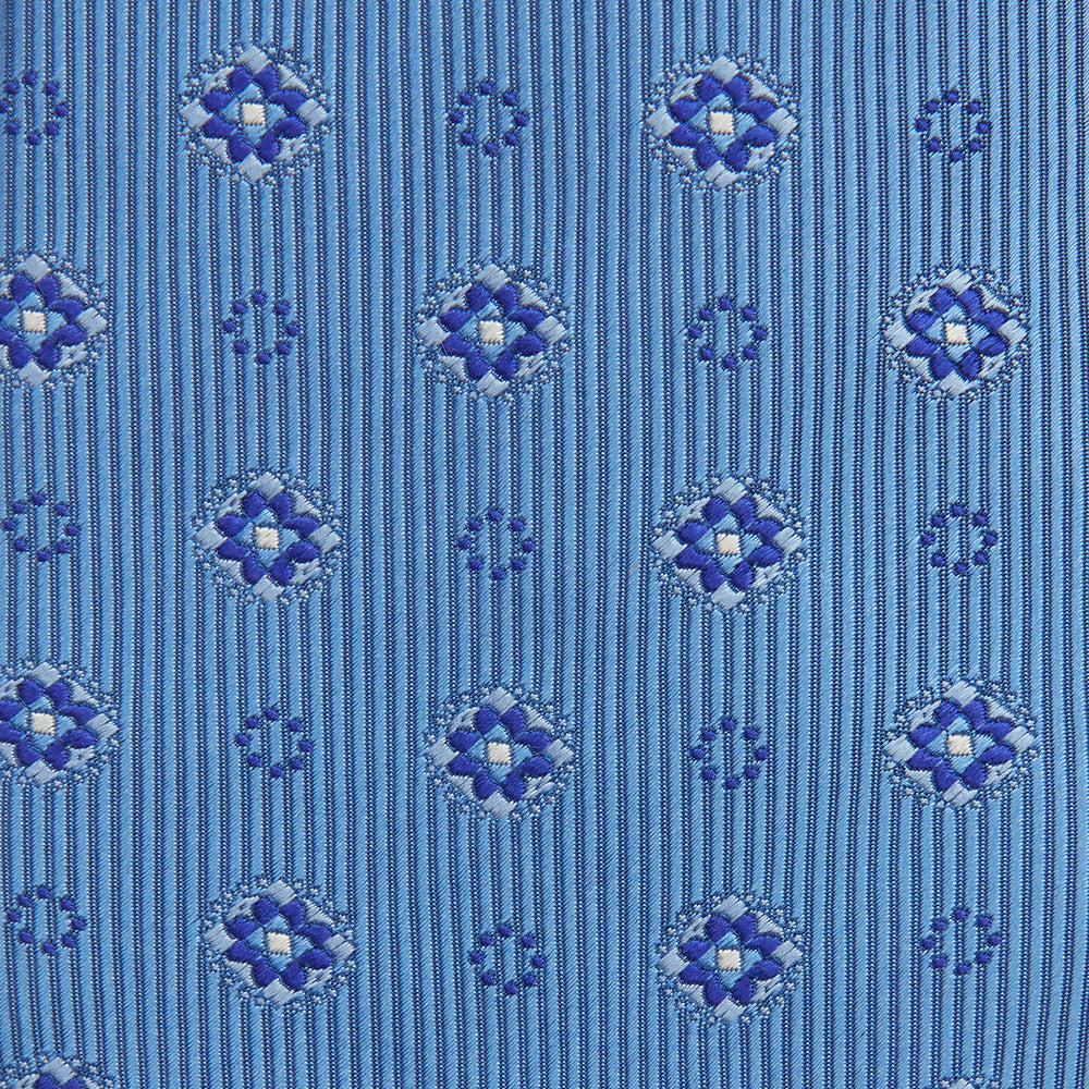 The Blue Multi Floral Motif Silk Tie by Turnbull & Asser features subtle diagonal stripes and blue and white floral medallions on light blue silk—ideal for bold shirts. The tie is shown rolled at one end.