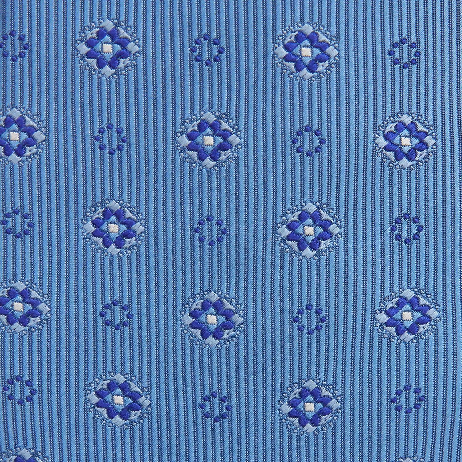 View 3 of product The Blue Multi Floral Motif Silk Tie by Turnbull & Asser features subtle diagonal stripes and blue and white floral medallions on light blue silk—ideal for bold shirts. The tie is shown rolled at one end.