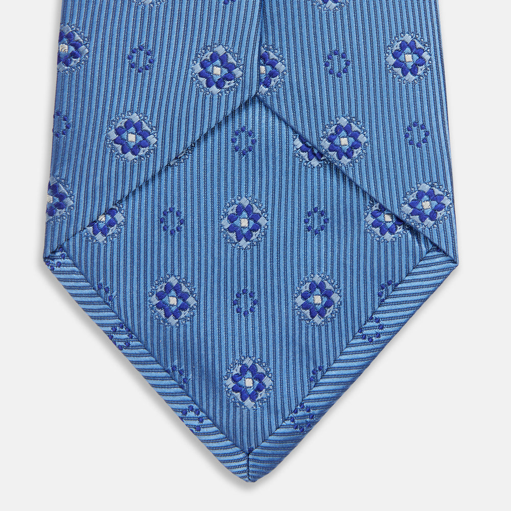 The Blue Multi Floral Motif Silk Tie by Turnbull & Asser features subtle diagonal stripes and blue and white floral medallions on light blue silk—ideal for bold shirts. The tie is shown rolled at one end.