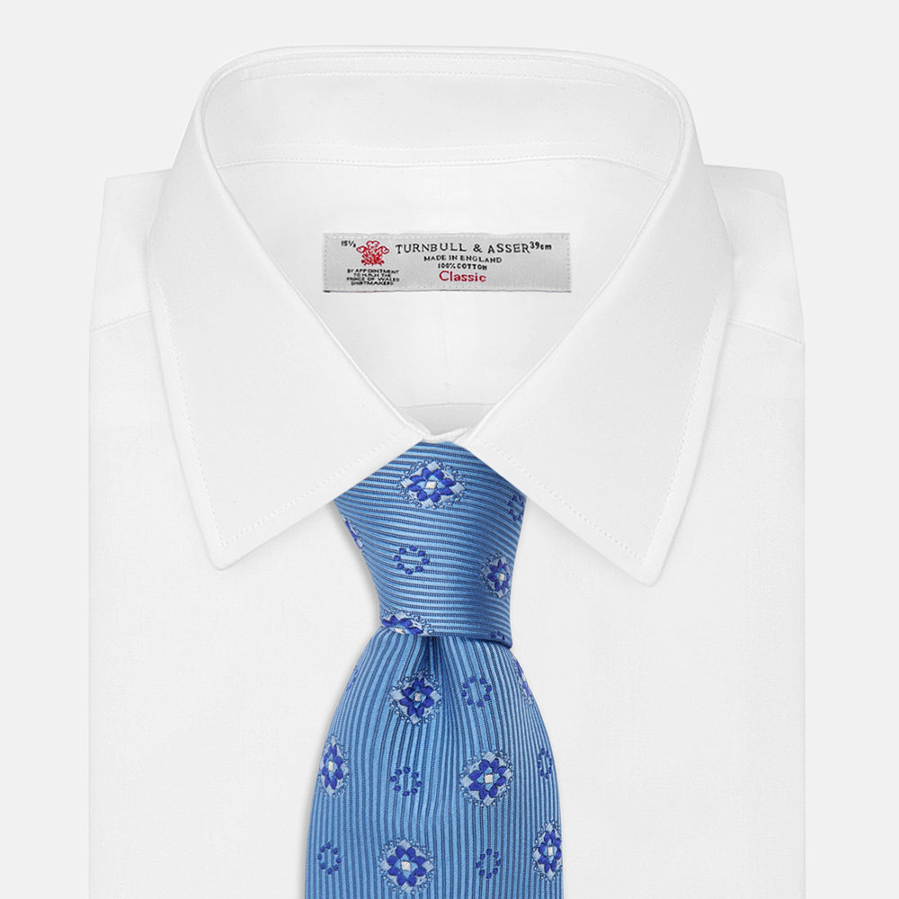 The Blue Multi Floral Motif Silk Tie by Turnbull & Asser features subtle diagonal stripes and blue and white floral medallions on light blue silk—ideal for bold shirts. The tie is shown rolled at one end.