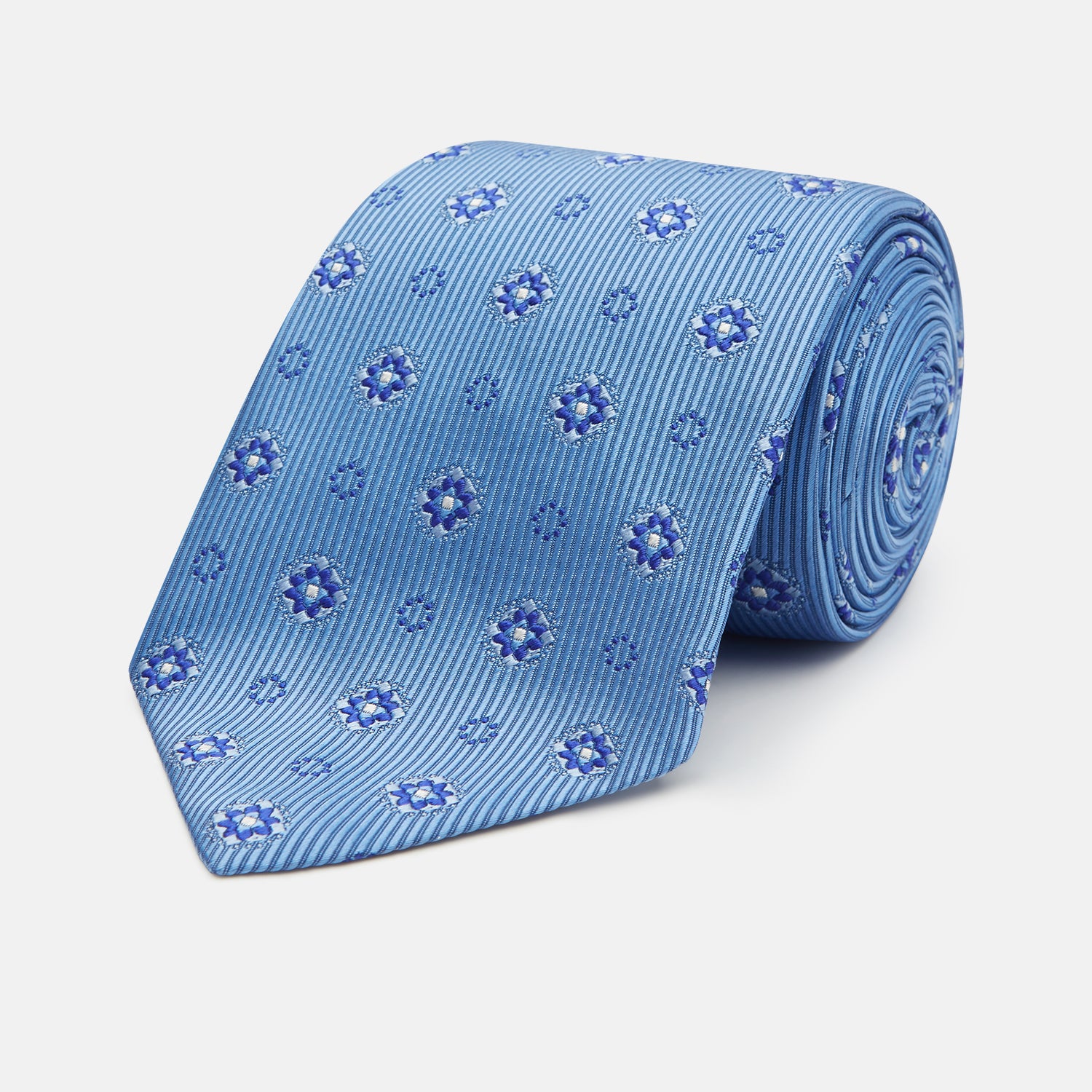 View 1 of product The Blue Multi Floral Motif Silk Tie by Turnbull & Asser features subtle diagonal stripes and blue and white floral medallions on light blue silk—ideal for bold shirts. The tie is shown rolled at one end.