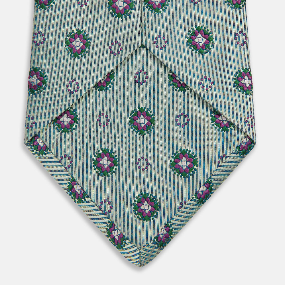 The Green Multi Floral Motif Silk Tie by Turnbull & Asser features light gray silk with thin diagonal stripes and a floral medallion pattern in pink, purple, and green—ideal for adding summer color to your outfit. Displayed rolled up on a white background.
