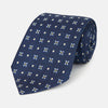 View 1 of product The Turnbull & Asser Navy Floral Emblem Tie displays a repeating pattern of small light blue, white, and yellow floral and diamond motifs, highlighting refined elegance, shown rolled up on a white background.