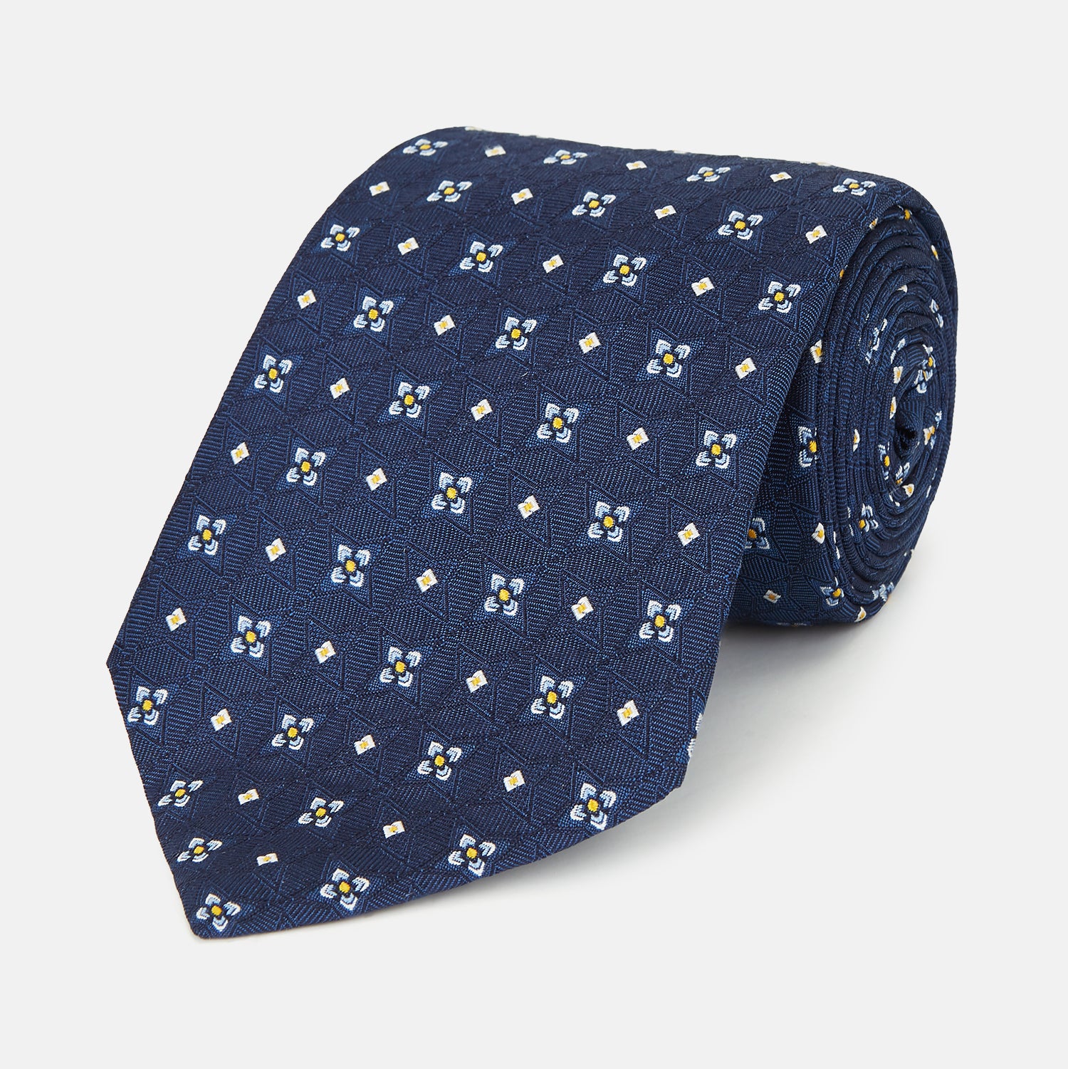 View 1 of product The Turnbull & Asser Navy Floral Emblem Tie displays a repeating pattern of small light blue, white, and yellow floral and diamond motifs, highlighting refined elegance, shown rolled up on a white background.